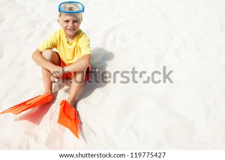 Young blonde boy sitting in white beach sand wearing his flippers and goggles as he waits to go swimming in the sea with copyspace