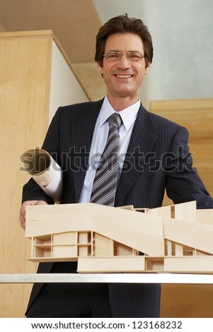 Smiling male architect with blueprints under his arm and holding a three dimensional building model
