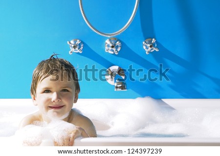 Smiling little boy peeking over the top of a frothy bubble bath against a blue wall