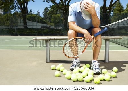 man, holding a tennis racquet, sitting on a bench holding his head down, tennis balls in front of him, tennis net visibile on his right side, trees in the background