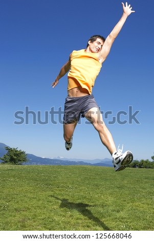 man jumping off the ground with left hand extended up, right hand at the back, blue skies visible overhead, green grass visible at the bottom