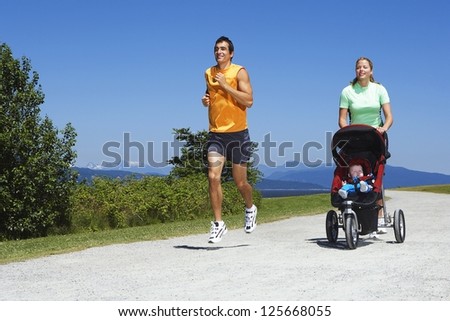 woman pushing a stroller with baby walking beside a man jogging, with bushes and trees in the background