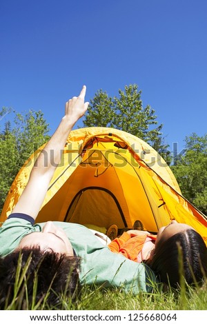 couple lying on the grass, their feet inside the tent in the background, sky and trees seen overhead