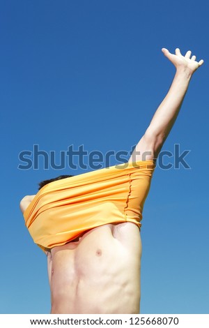 man trying to wear an orange shirt, right arm extended up, blue sky visible overhead