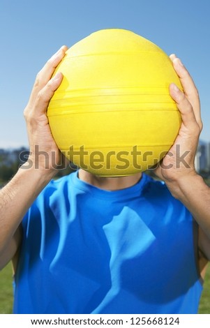man holding up a yellow ball up covering his face, blue sky seen ion the background