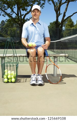 man sitting on a bench holding tennis balls with his left hand, tennis racket leaning against the bench, net visible on his right side, trees in the background