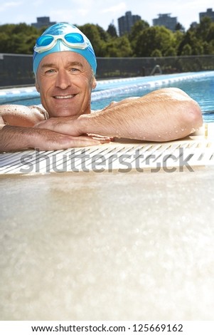 smiling man in a swimming wearing swimming cap and goggles on his head, his arms resting at the edge of swimming pool