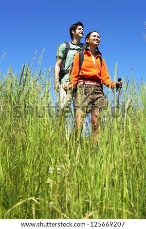 couple hiking together, blades of grass in the foreground, both wearing backpacks and holding hiking stick