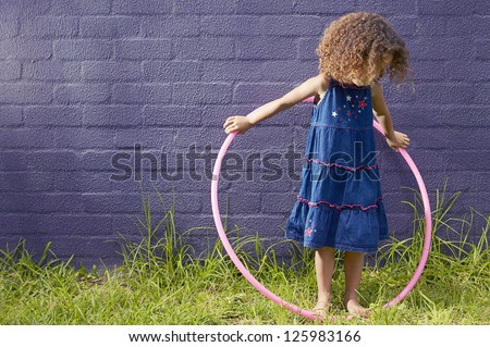 Little girl with curly hair standing in front of a blue brick wall looking down at her hula hoop which she is holding