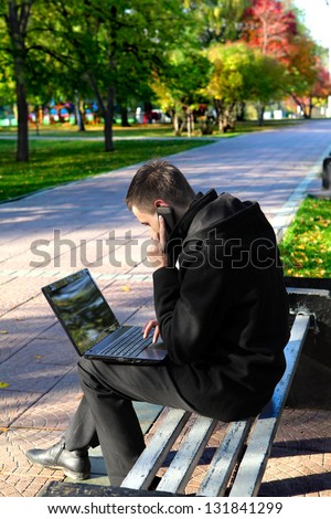 Busy Young man with Laptop sitting on the bench at the Autumn Park and Talking on Mobile Phone