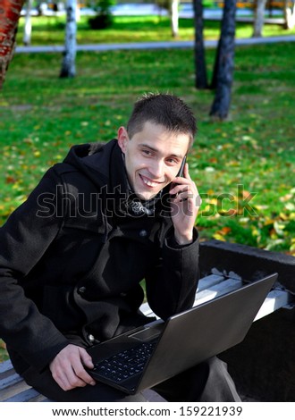 Happy Man with Laptop Taking on Cellphone on the bench at the Autumn Park