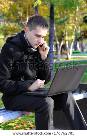 Worried Young man with Laptop sitting on the bench at the Autumn Park and Talking on Mobile Phone