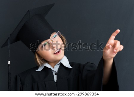 Cute pupil in mortar board pointing at something at the black chalkboard in classroom.