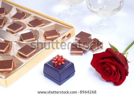 Valentine table with rose, box of chocolates, gift box and two glasses of white wine on white background.