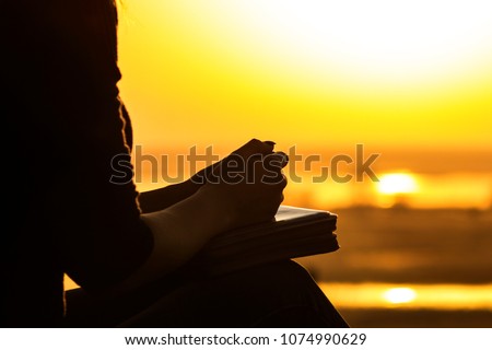 silhouette of the hands of a young woman praying to God in the nature, the girl holds her hands on the Bible near the tree at sunset, the concept of religion and spirituality