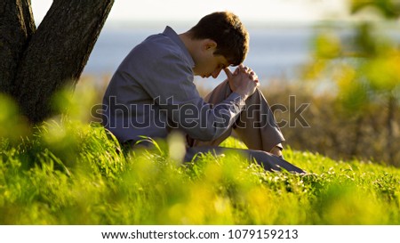young man praying to God near a tree in the nature bowing his head to his knees with gratitude, male asks for help finding solace in faith, concept religion