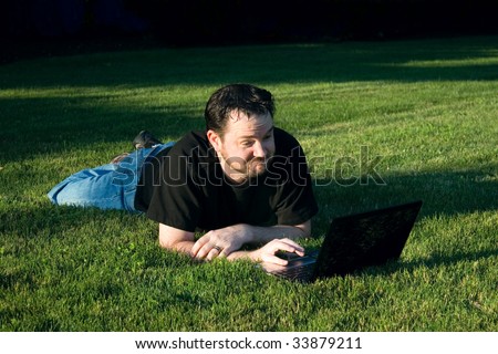 A young Caucasian man has an interesting smirk on his face as he speaks to the girl on his webcam on the lush green grass.