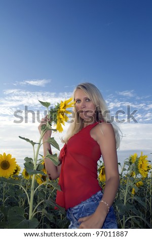 Beautiful girl in a red dress and jeans holds in a hand a sunflower in a field. Summer evening. Blue sky and white clouds above sunflower field