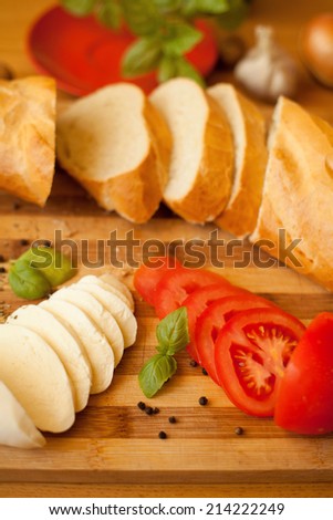 Mozzarella cheese with fresh tomatoes and baguette. Cut mozzarella cheese and cut fresh tomatoes are surrounded by fresh baguette. Everything is presented on wooden bread- board.