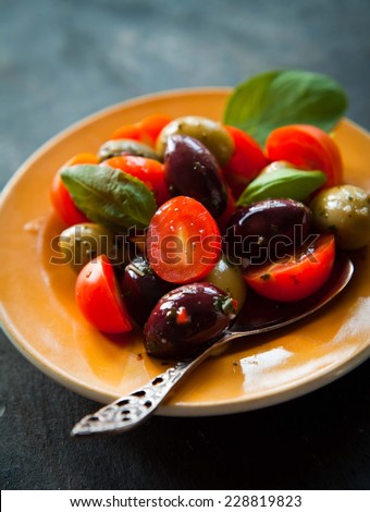 Green and black olives with small, cherry tomatoes mixed with oil and herbs on a yellow plate. Decorated with basil leaves. Vegetables popular in Italy.