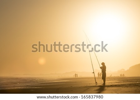 Man Fishing In The Sea At The Sunset, Scenic View, Sand Sun And Waves In The Water. Figure Silhouette.