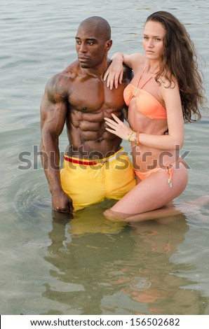 Fit happy couple enjoying their vacation at the beach, a strong black man and a white brunette sexy woman. Woman in swimsuit holding her hand on his perfect abs