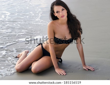 Sexy brunette girl in black swimsuit relaxing on the beach while waves hit her hot fit body