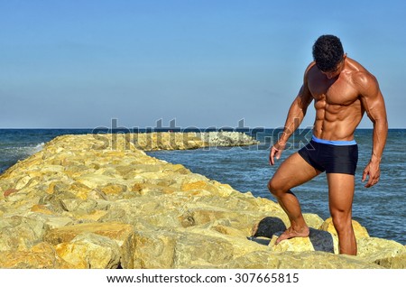 Strong bodybuilder with six pack.Fitness trainer with perfect abs, shoulders,biceps, triceps,chest, flexing his muscles on the beach with sea waves on the background, training in vacation