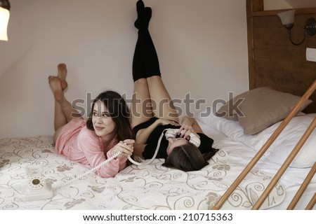 image of a two girls on the bed happy talking on phone dreamy beauty girls stretching her legs while lying in bed against the wooden wall