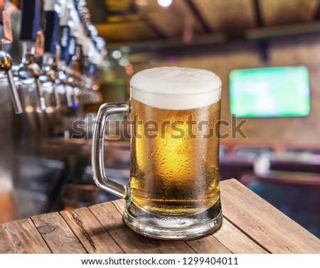 Frosty mug of light beer on the old wooden table. Pub interior and bar counter with beer taps at the background.