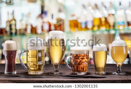 Cold mugs and glasses of beer on the old wooden table. Pub interior and bar counter with beer taps at the background. Assortment of beer.