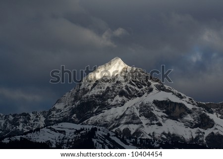 A sunbeam illuminates the peak of a tall mountain in the alps, as a darkness approaches