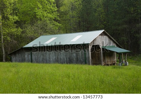 Side view of a weathered old pole barn, with green, roof, in a pasture, in front of a forest.