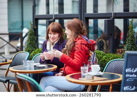 Two cheerful young girls spending time together and chatting in a Parisian street cafe on a spring or fall day