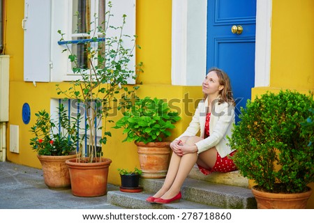 Beautiful young woman in red polka dot dress sitting on a house porch on a Parisian street with colorful bright houses