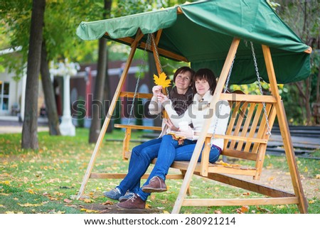 Middle aged woman with her adult daughter on a swing on a fall day, holding autumn leaves in their hands