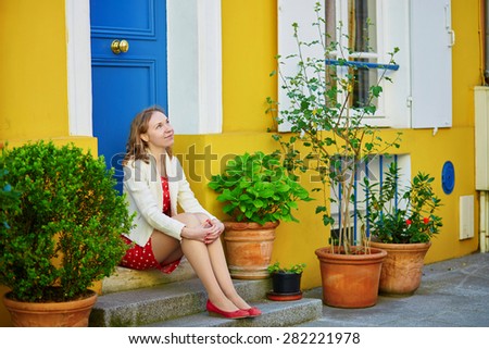 Beautiful young woman in red polka dot dress sitting on a house porch on a Parisian street with colorful bright houses