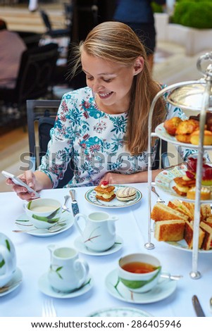Beautiful young woman enjoying afternoon tea with selection of fancy cakes and sandwiches in a luxury Parisian restaurant. Girl is checking her mobile phone