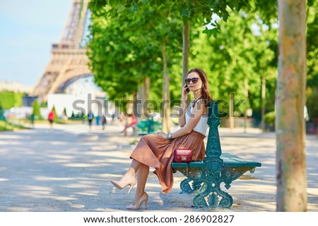 Beautiful young Parisian woman in long skirt near the Eiffel tower on a summer day, sitting on the bench and speaking on mobile phone