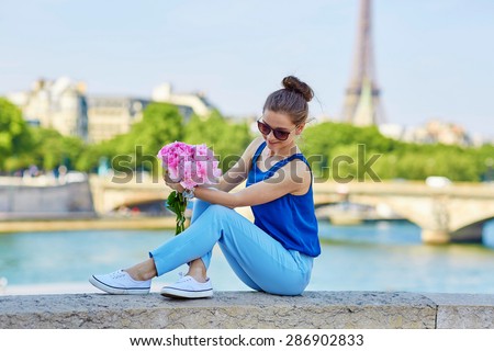 Beautiful young Parisian woman in blue tube top with bunch of pink peonies on the bank of the Seine, Eiffel tower is in the background