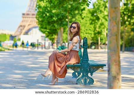 Beautiful young Parisian woman in long skirt near the Eiffel tower on a summer day, sitting on the bench