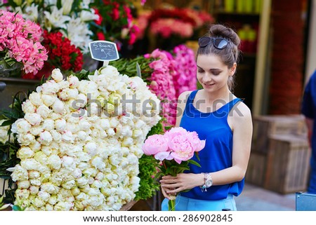 Beautiful young Parisian woman in blue tube top selecting pink peonies in French flower shop or at market