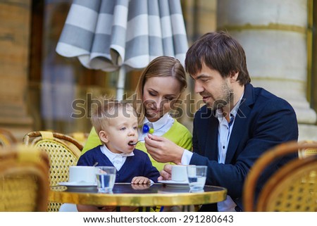 Happy family of three spending fun together in an outdoor Parisian cafe, father is feeding his son