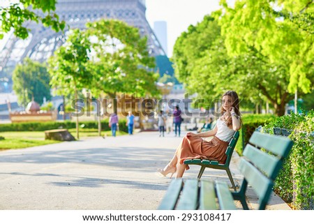 Beautiful young Parisian woman in long skirt sitting on the bench near the Eiffel tower on a summer day