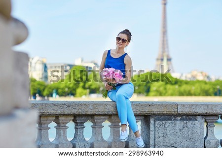 Beautiful young Parisian woman in blue blouse with bunch of pink peonies on the Alexandre III bridge in Paris, Eiffel tower is in the background