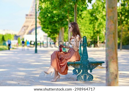 Beautiful young Parisian woman in long skirt near the Eiffel tower on a summer day, sitting on the bench and using mobile phone