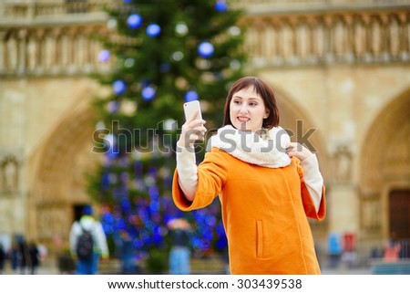 Happy young tourist in Paris on a winter day, taking selfie with her mobile phone, main Parisian Christmas tree and Notre-Dame cathedral in the background