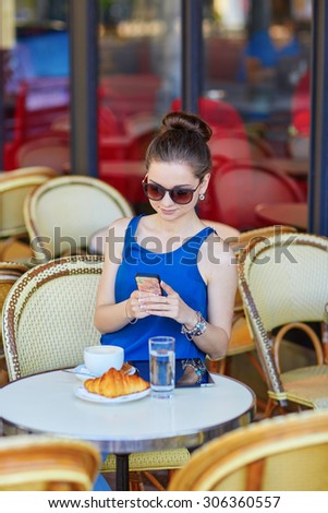 Beautiful young Parisian woman in blue blouse drinking coffee and and taking photo with her mobile phone in an outdoor cafe on a summer day