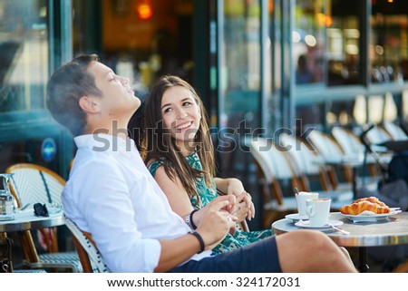 Young romantic couple drinking coffee, eating traditional French croissants and smoking in a cozy outdoor cafe in Paris, France