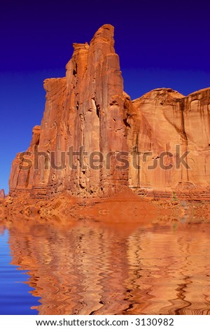 A digital composite red-orange cliff reflected in gently rippling water under deep blue skies.